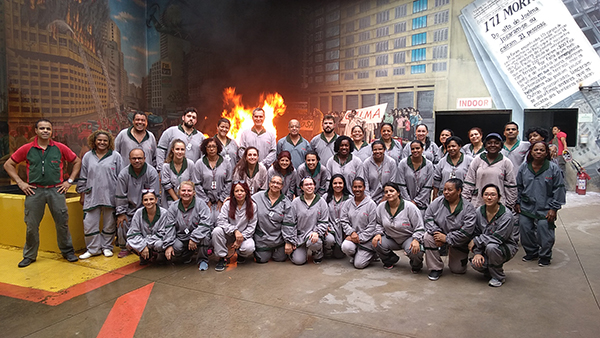 Foto de uma turma de servidores em treinamento, todos de roupa cinza e com um chama ao fundo. 
