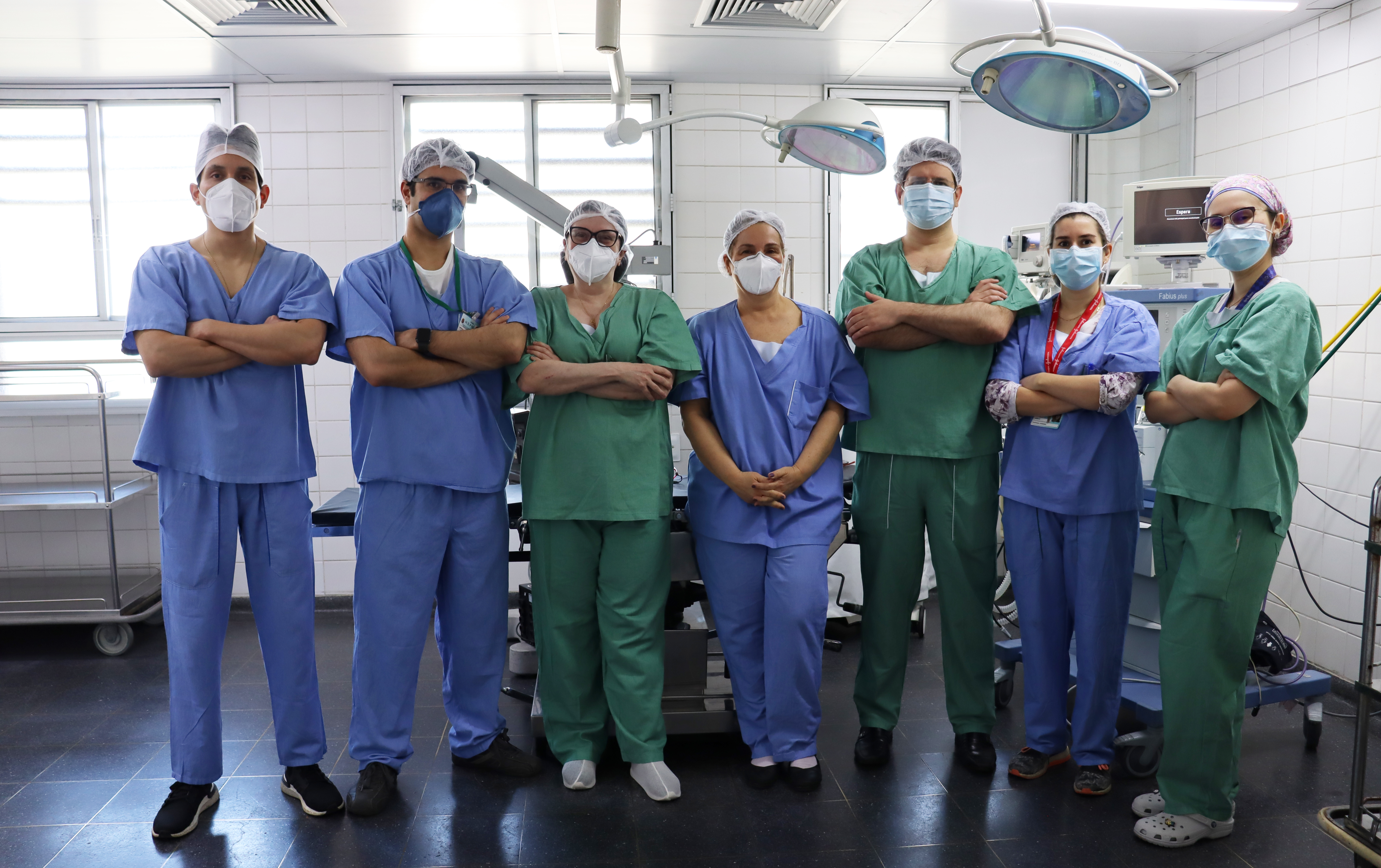 Foto da equipe de cabeça e pescoço, composta por sete pessoas, todos uniformizados e fazendo pose em uma sala de cirurgia. 