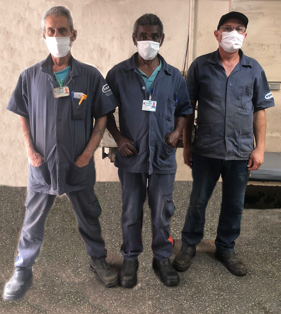 Foto dos 3 encanadores do hospital, em frente a uma parede bege e usando macacão azul e máscara branca