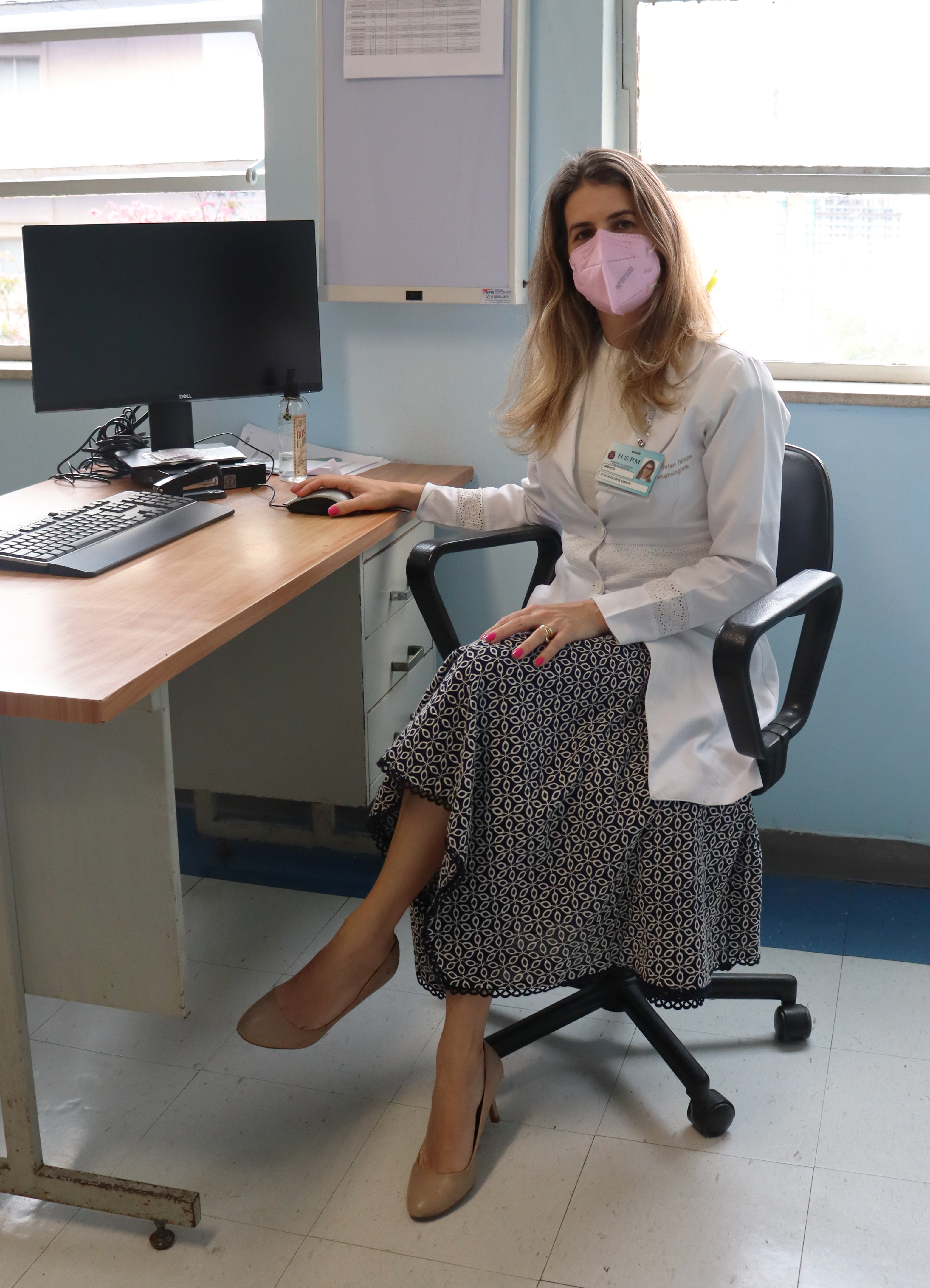 Foto da médica, sentada no consultório sentada em uma cadeira em frente a uma mesa com computador. 