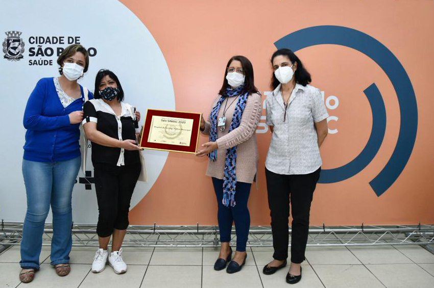 Foto das funcionárias recebendo a placa do prêmio Sinasc, duas mulheres do lado esquerdo e duas do lado direito
