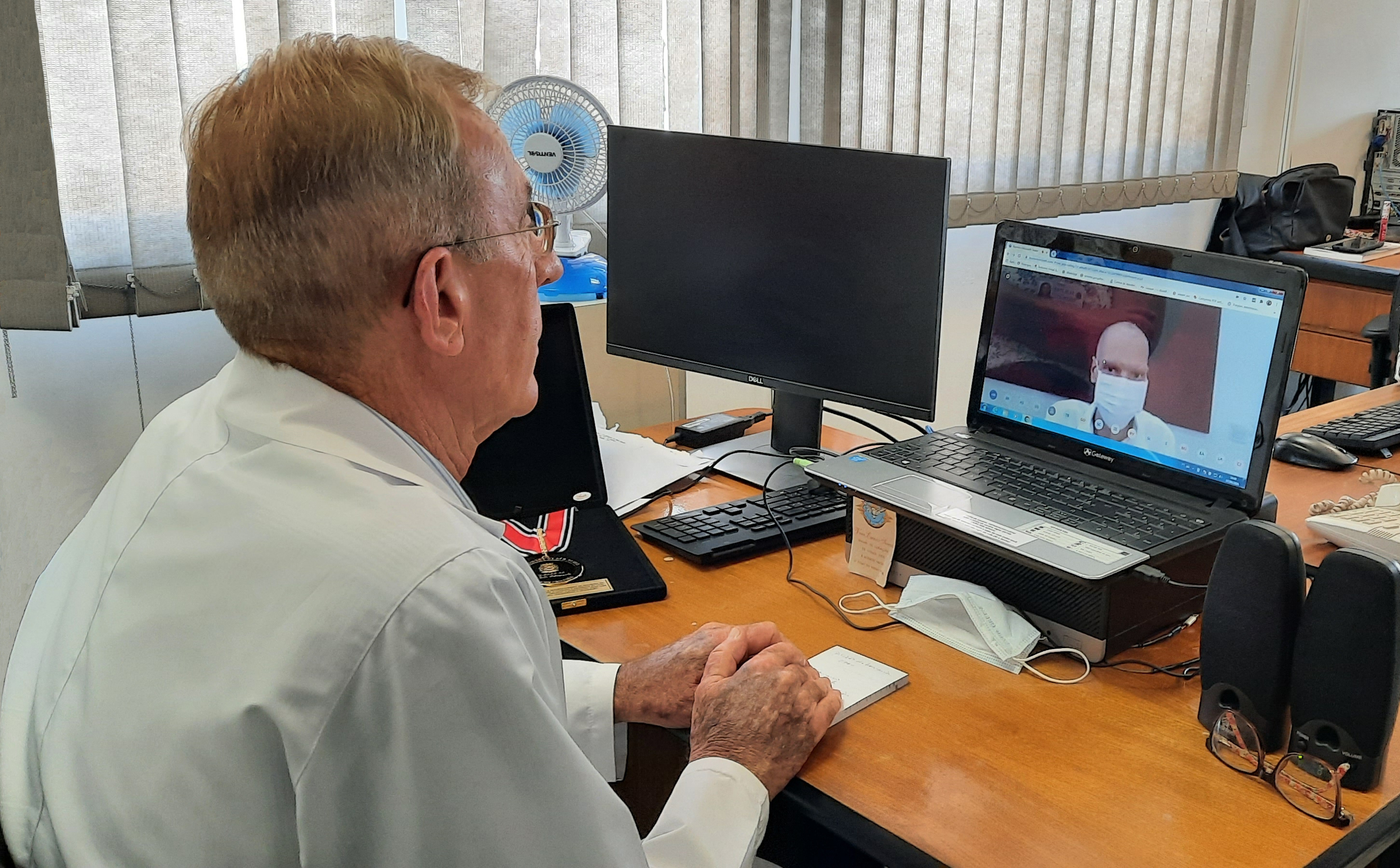 dr. Volpiani sentado em frente ao computador que transmitiu a cerimônia. na tela, aparece o prefeito Bruno Covas de máscara branca.