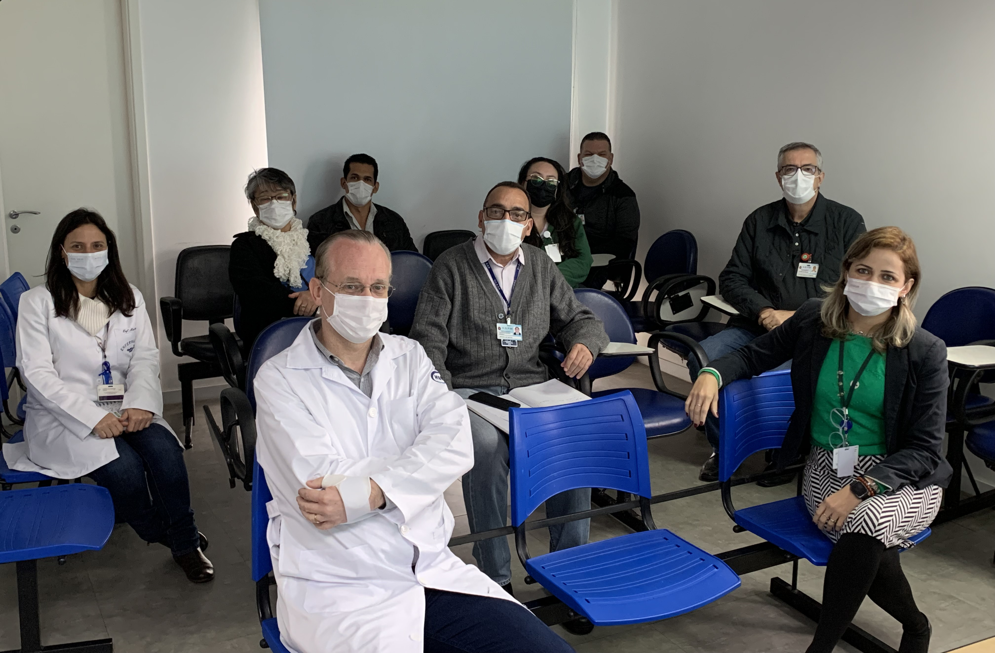 Foto de nove membros da comissão, sentados em uma sala branca com cadeiras azuis