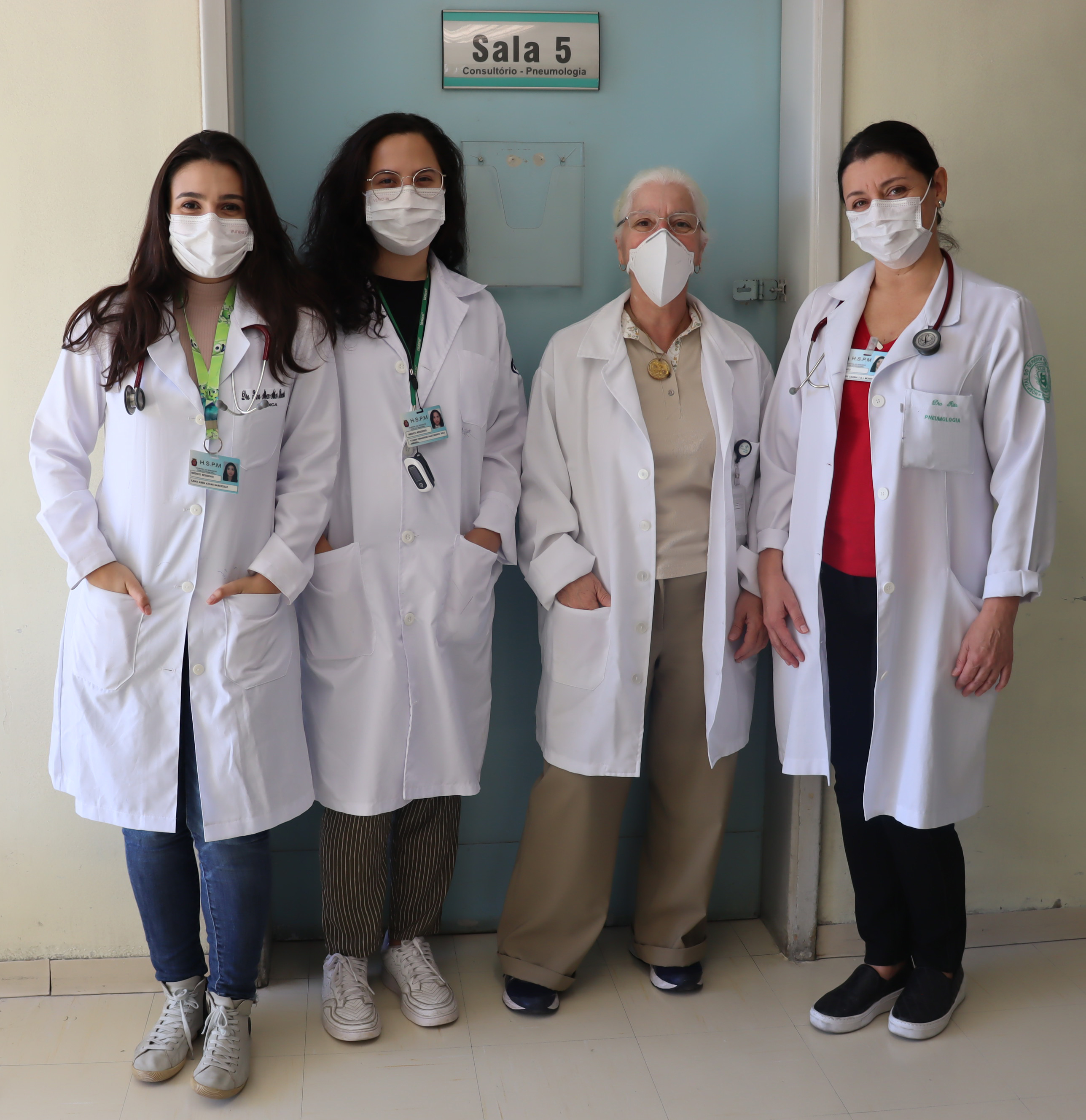 Foto com parte da equipe de pneumologista, quatro mulheres, em frente a uma porta azul onde está escrito sala cinco consultório pneumologia