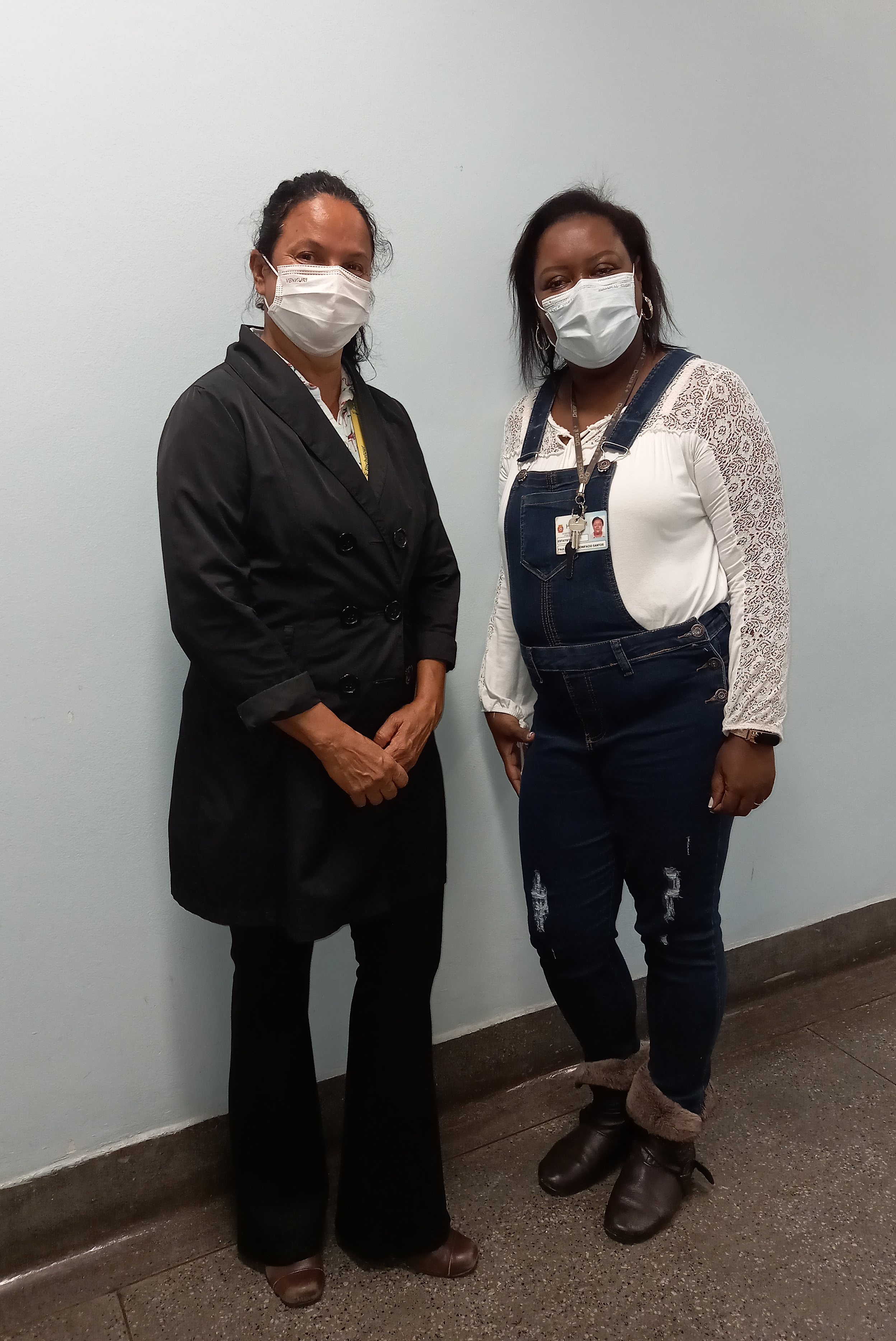 Foto de duas mulheres morenas em pé, sorrindo, em frente a uma parede branca