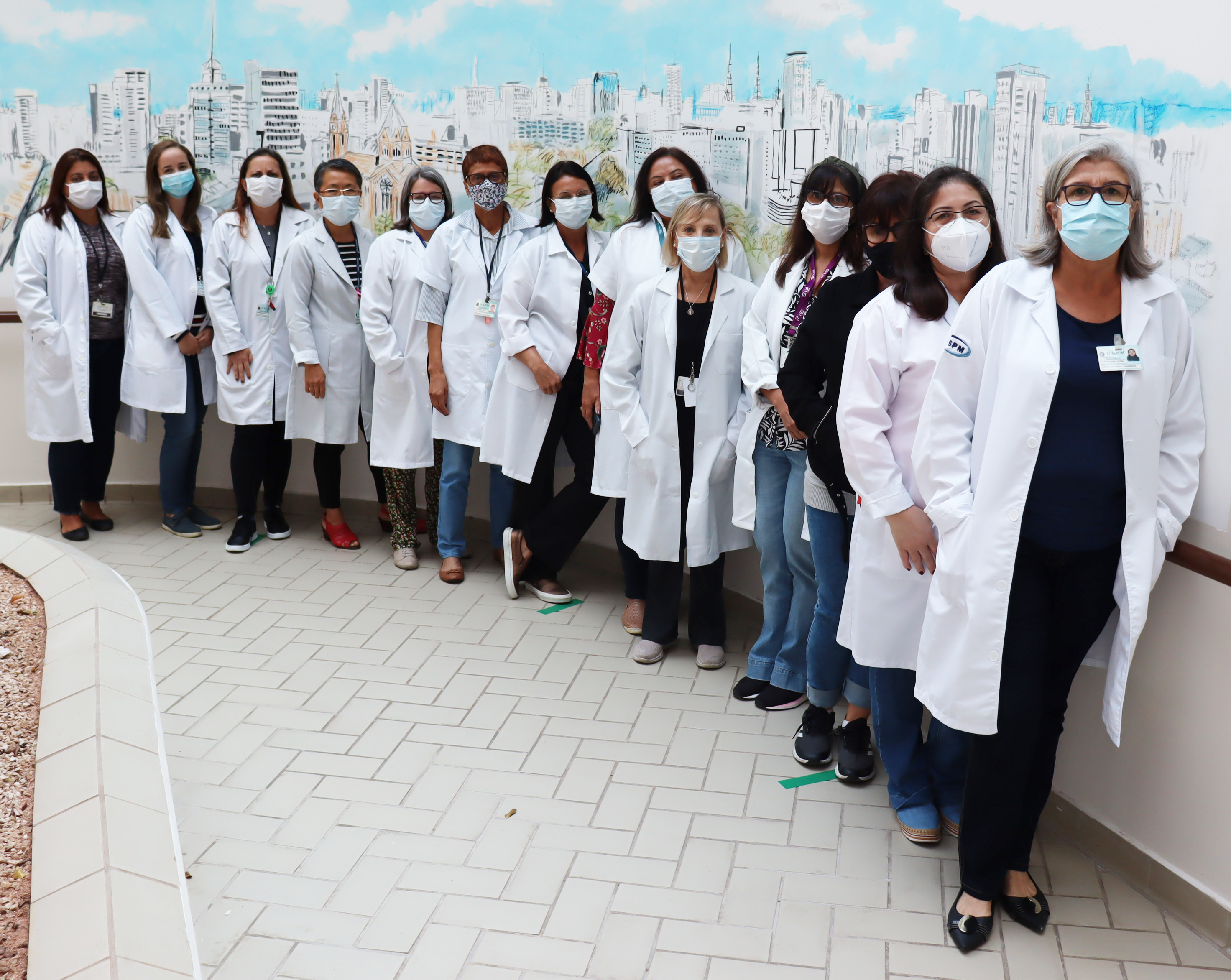 Foto com 13 assistentes sociais do HSPM, todas de jaleco branco,  com um grafite da cidade de São Paulo ao fundo.
