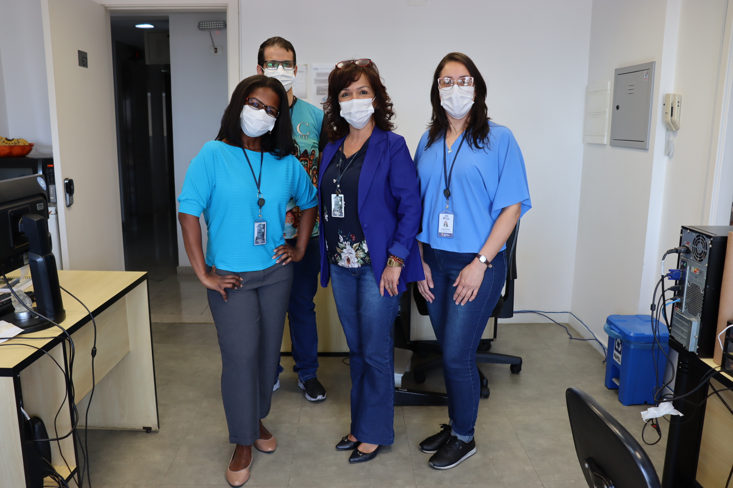 Foto de 4 funcionários juntos de pé, dentro da sala de trabalho. Todos vestem roupa azul