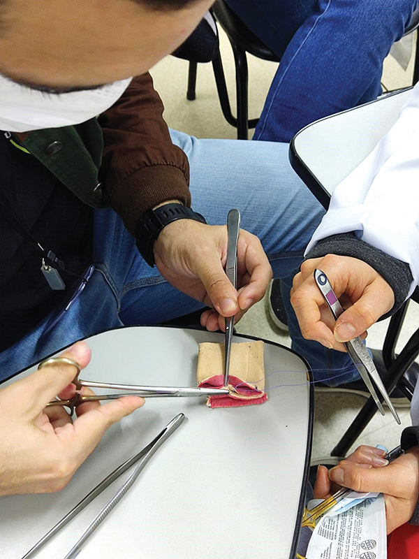 Imagem das mãos de três alunos que simulam uma sutura