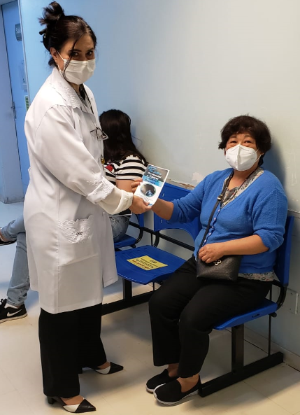 Imagem da médica em pé entregando um folder informativo para a paciente que está sentada