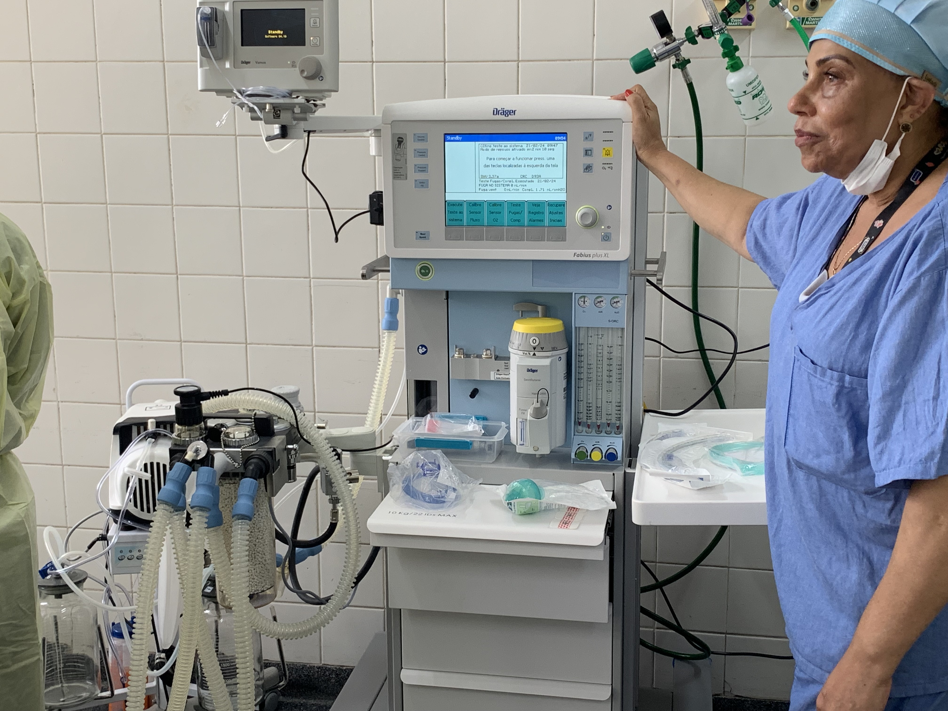 Fotografia do aparelho de anestesia no centro cirúrgico. Ao lado está uma enfermeira , que toca no aparelho enquanto olha em outra direção.