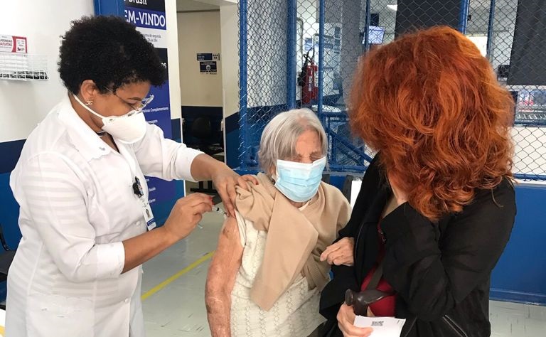 Foto de vacinação. À esquerda da imagem, uma profissional de saúde de jaleco branco está aplicando a vacina em uma idosa mais baixa, ao seu lado, de cabelo grisalho e máscara de proteção azul. A senhora está segurando o braço de uma mulher mais jovem, de cabelos ruivos e roupa preta, que está ao seu lado.
