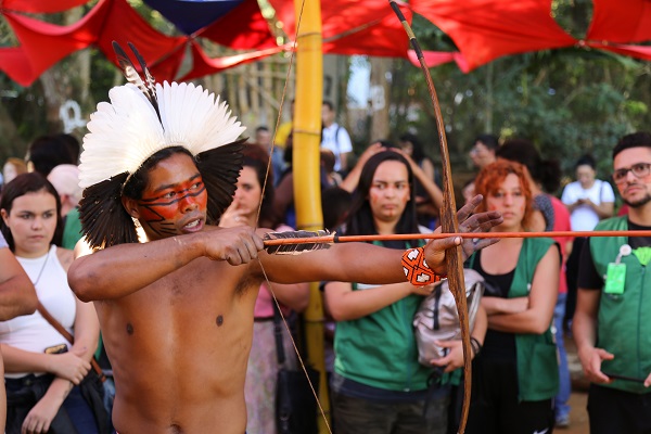 #PraCegoVer: na imagem há um indígena com pintura na pele, cocar e um arco e flecha nas mãos. Ao fundo há profissionais do Programa Ambientes Verdes e Saudáveis (PAVS)