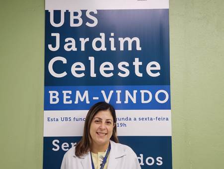 #PraCegoVer: na foto, Ingrid está de jaleco branco sorrindo. Atrás dela há um cartaz de fundo azul . Em letras brancas está escrito UBS Jardim Celeste. Bem-vindo