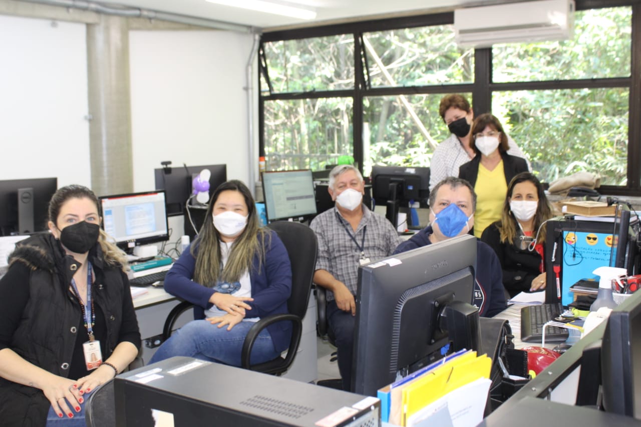 Em uma sala cheia de computadores estão cinco mulheres e dois homens. Todos usam máscara de proteção.