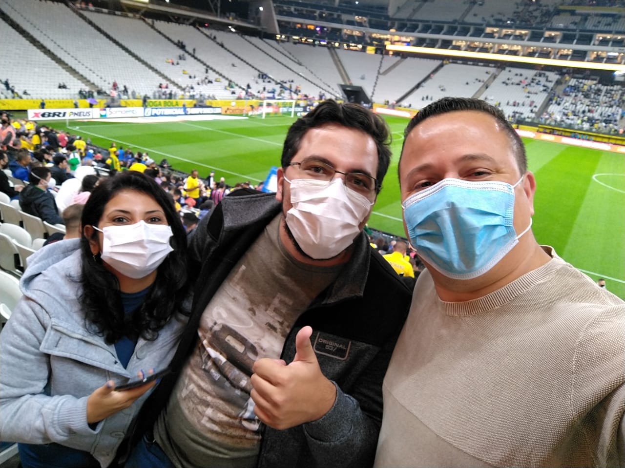 Dois homens e uma mulher estão usando máscara de proteção na arquibancada de um estádio. Atrás deles, está um campo de futebol. O homem que está no meio está fazendo um sinal positivo com a mão esquerda.