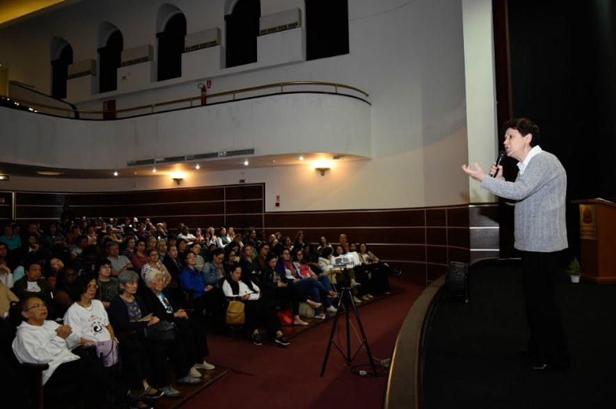 Lia Diskin Palas Athena fala ao microfone no palco durante Encontro de 15 anos de Práticas Integrativas e Complementares em Saúde no Colégio Caetano de Campos. Há dezenas de pessoas na plateia.