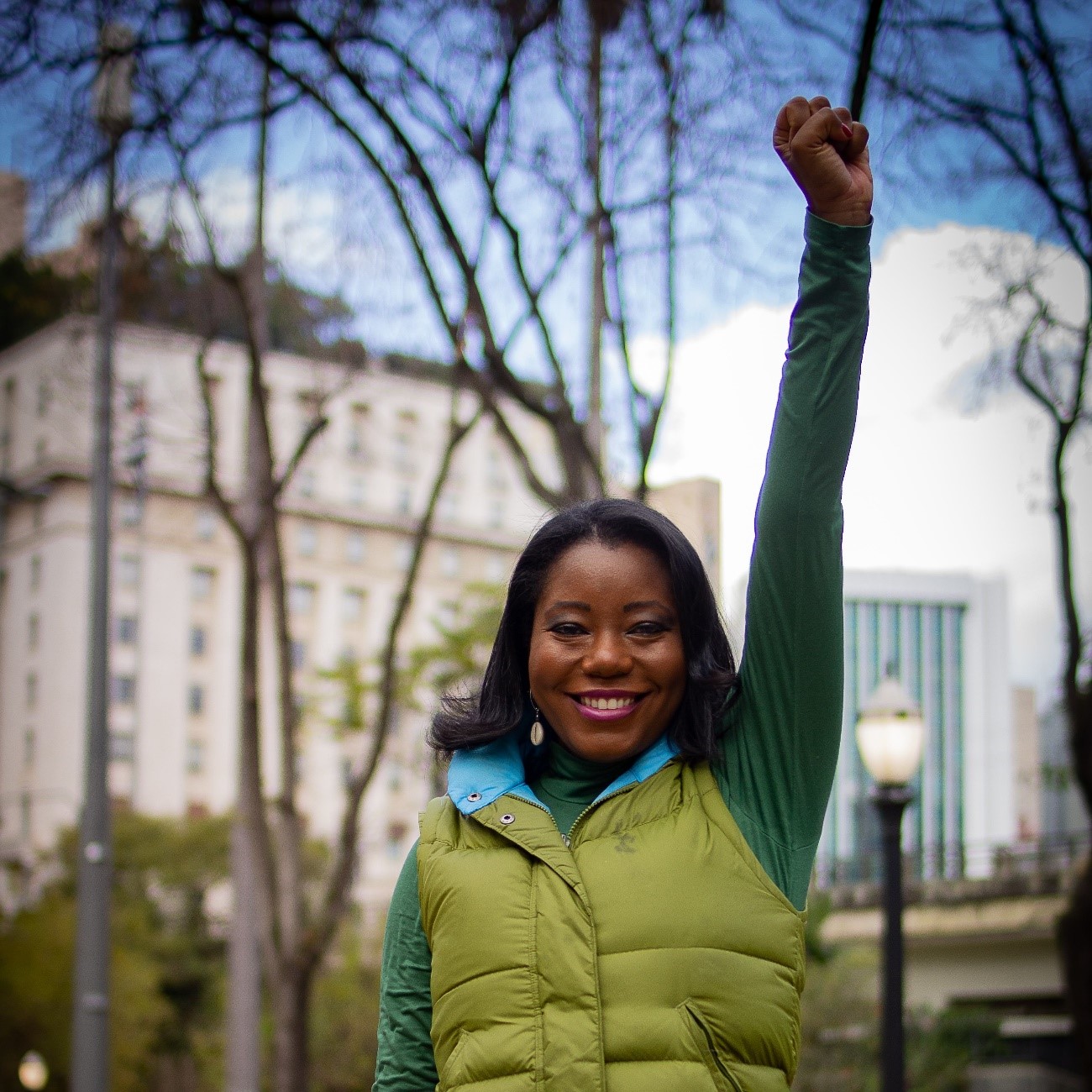 A médica Lígia Santos está sorrindo e com um dos punhos erguidos, em símbolo de luta. Ela usa um colete verde claro em cima de uma blusa verde escuro de mangas longas. A gola do colete é azul claro.