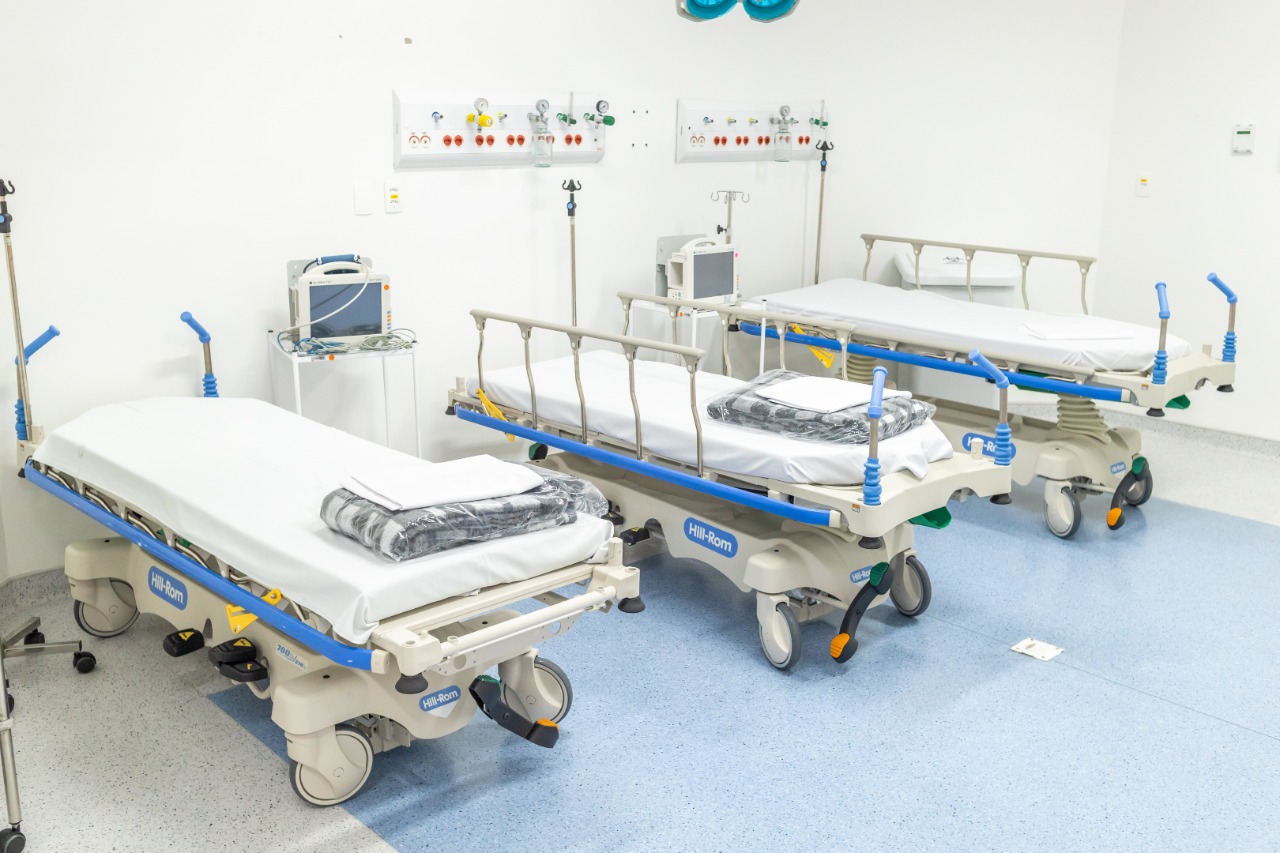 Na imagem há três macas claras com detalhes em azul. Em cima de cada uma delas há um lençol dobrado. Nesta sala também tem equipamentos de saúde.