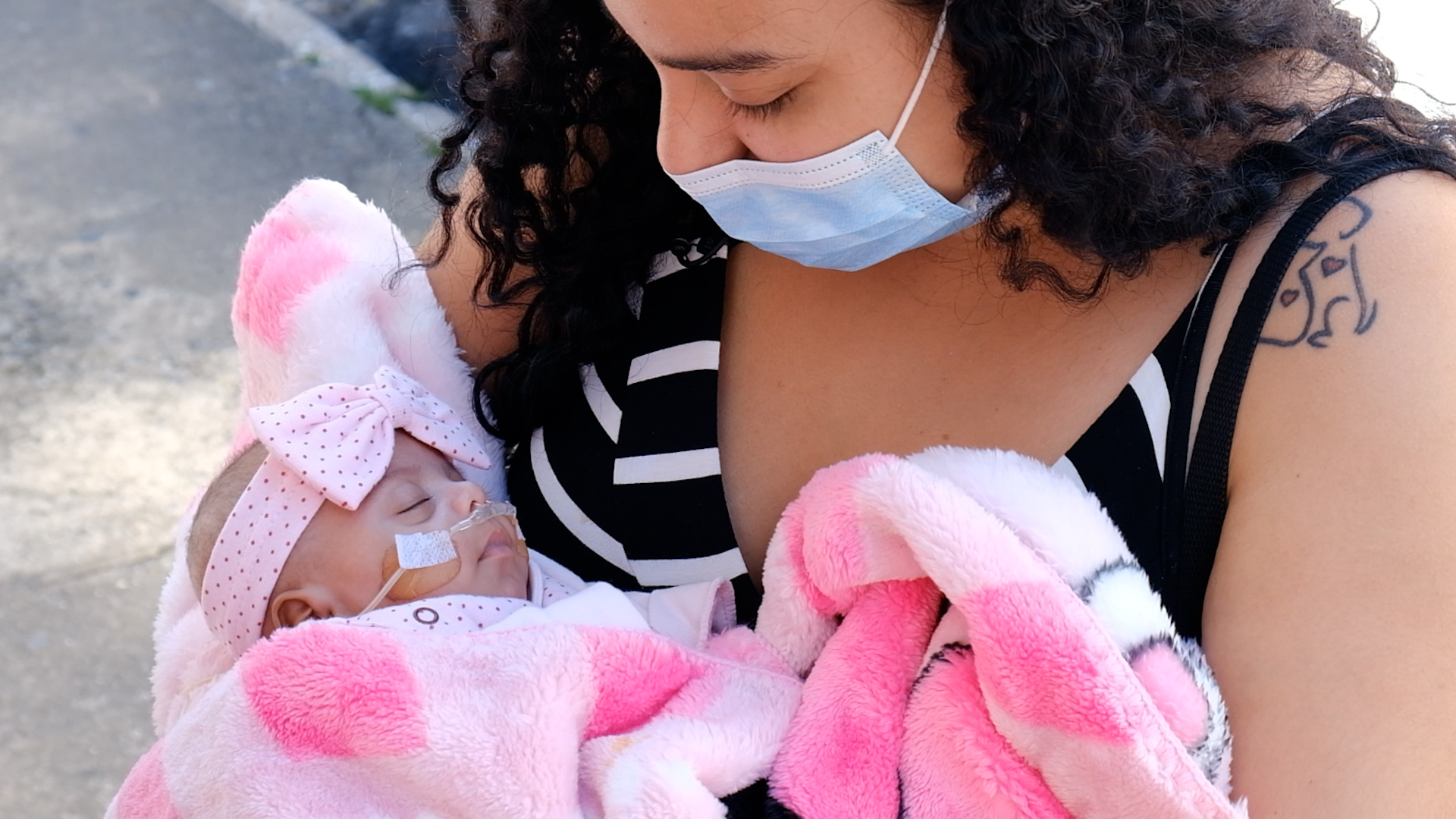 A foto mostra Camila, uma mulher jovem de cabelos escuros, segurando nos braços um pequeno bebê envolto em um cobertor cor-de-rosa