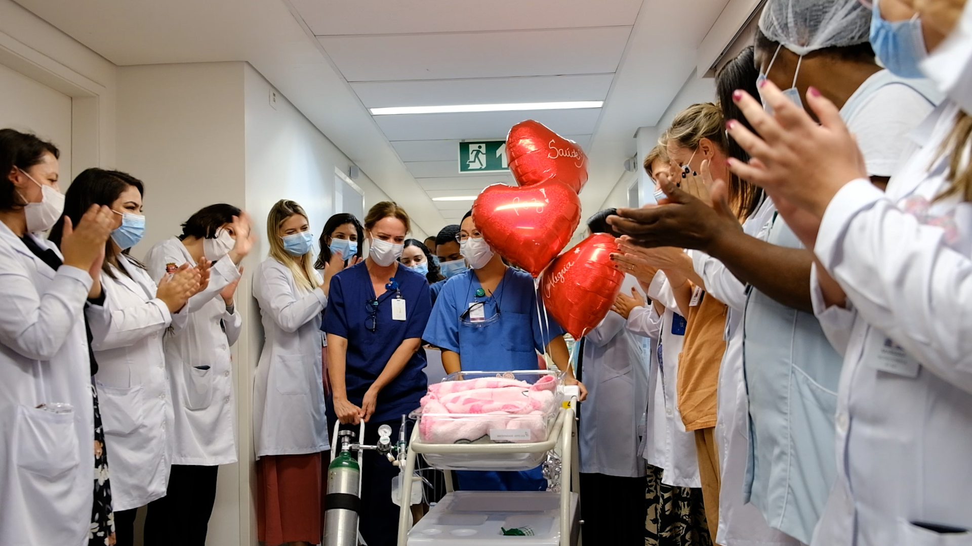 A foto mostra um ambiente hospitalar no qual mais de uma dezena de pessoas usando jalecos e máscaras aplaudem; todos estão voltados para um carrinho com um bebê, envolto em uma manta cor-de-rosa 
