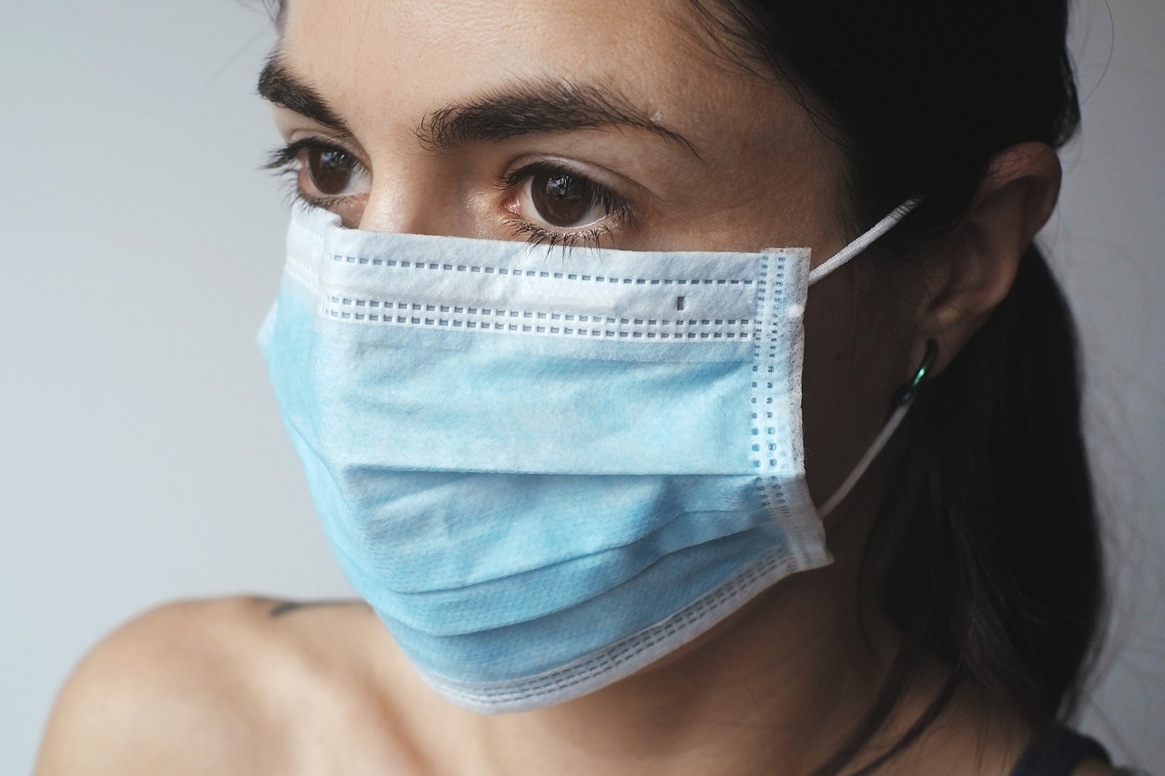 Uma mulher com cabelos pretos amarrados em um rabo de cavalo está usando uma máscara de proteção descartável, nas cores azul e branco.