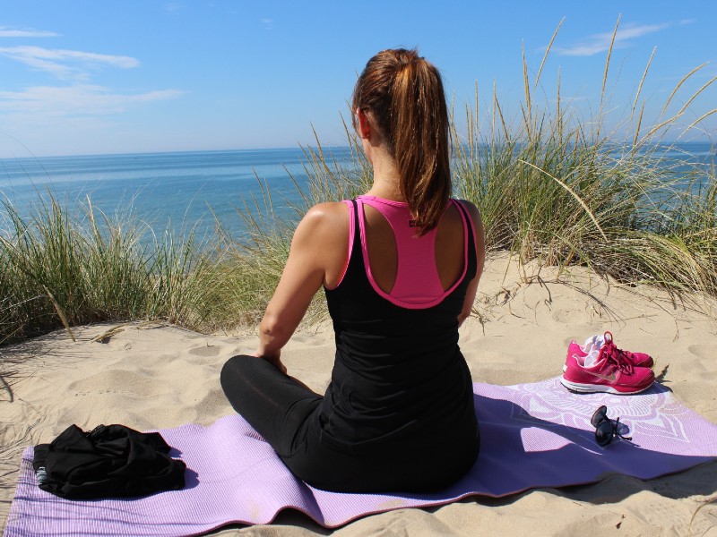 Um mulher usando regata e calça preta está sentada na areia sobre uma esteira de frente para o mar em posição de meditação. Há uma leve vegetação à frente e sobre a esteira há um óculos de sol, um par de tênis e um pano preto
