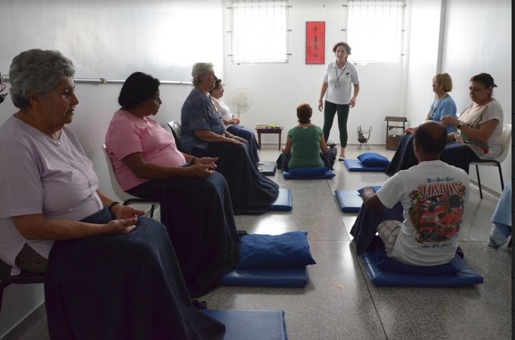 Um grupo de pessoas medita em uma sala. Alguns estão sentados em cadeiras, outros estão em colchonetes no chão.