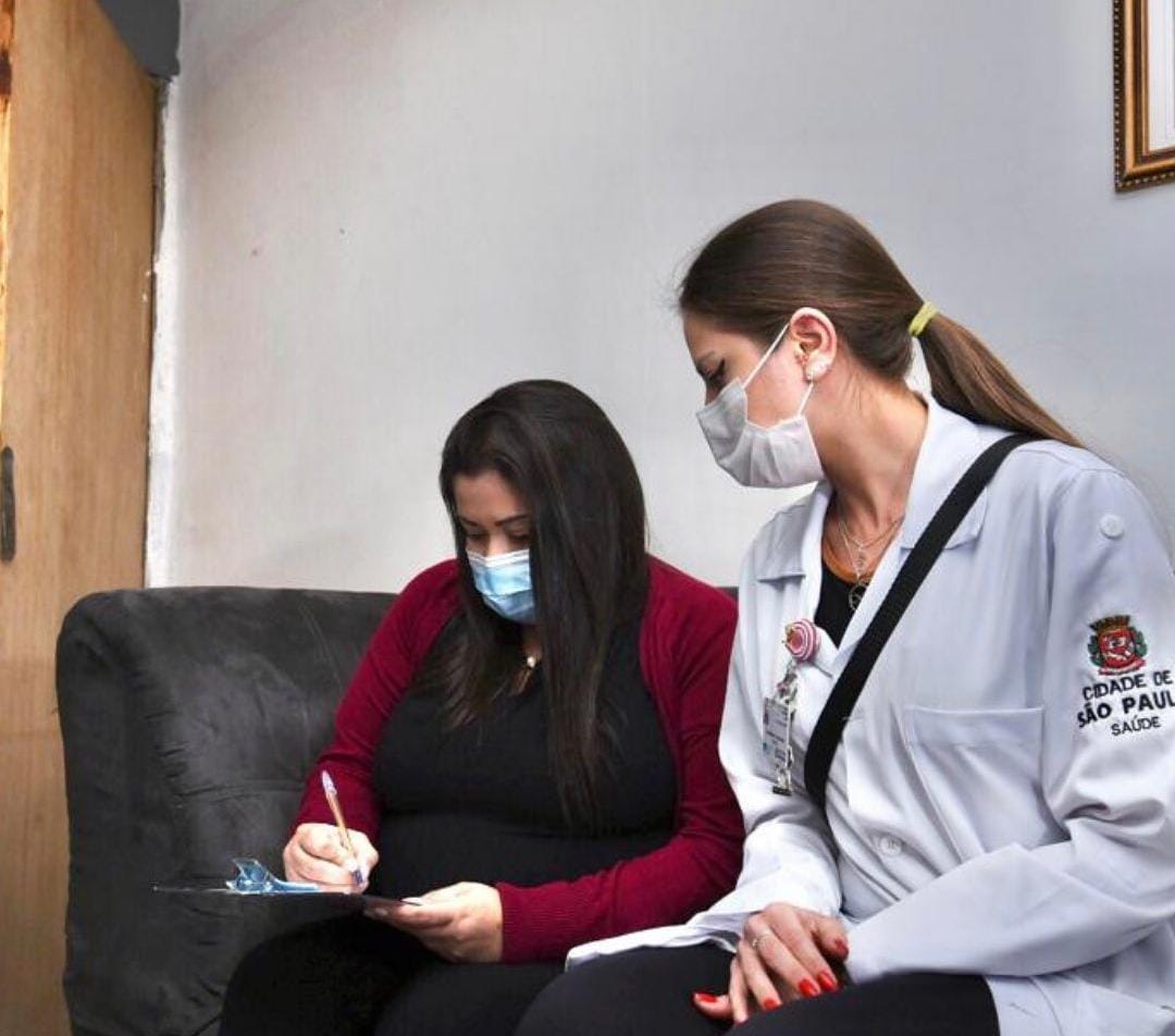 Duas mulheres estão sentadas em um sofá preto. A parede atrás é branca. À direita da imagem, uma das mulheres sentadas é uma profissional de saúde vestindo um jaleco branco e máscara de proteção branca. Ela está olhando para uma ficha que a outra mulher, sentada ao seu lado, está assinando. A mulher veste uma blusa preta, calça preta e uma blusa de frio vermelha aberta.