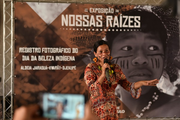 #PraCegoVer: Na imagem, fotografia de uma palestrante indígena, fazendo discurso. Ao fundo um poster com o tema principal dizendo: Exposição novas raízes.