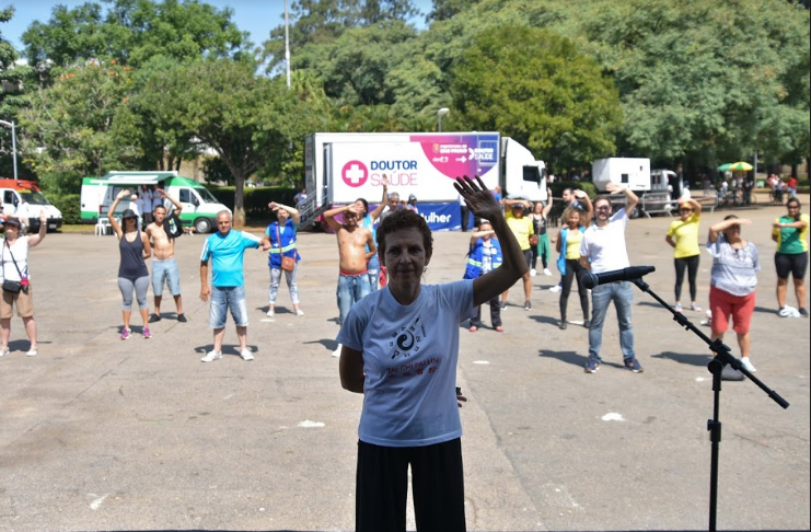 Um profissional orienta os movimentos de Práticas Integrativas para algumas pessoas no Parque Ibirapeura. O dia está ensolarado.