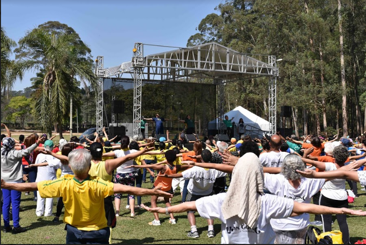 Dezenas de pessoas estão no gramado do Parque do Carmo fazendo atividade de Práticas Integrativas. ao fundo, num palco, três profissionais orientam os movimentos.
