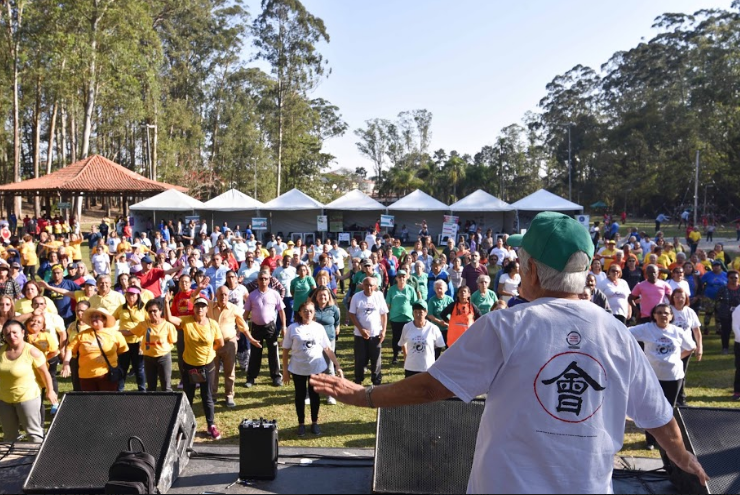  A foto foi tirada de cima do palco. Sobre o palco um profissional orienta os movimentos para dezenas de pessoas no gramado do Parque do Carmo. O dia está ensolarado.