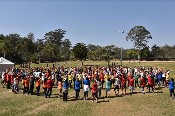 Dezenas de pessoas estão no gramado do Parque do Carmo fazendo atividade de Práticas Integrativas. O dia está ensolarado.