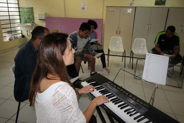 PraCegoVer: numa sala do Centro de Convivência e Cooperativa (Cecco) Santo Amaro, os pacientes estão sentados, cada um com uma folha de papel nas mãos, enquanto a musicista toca ao teclado musical.
