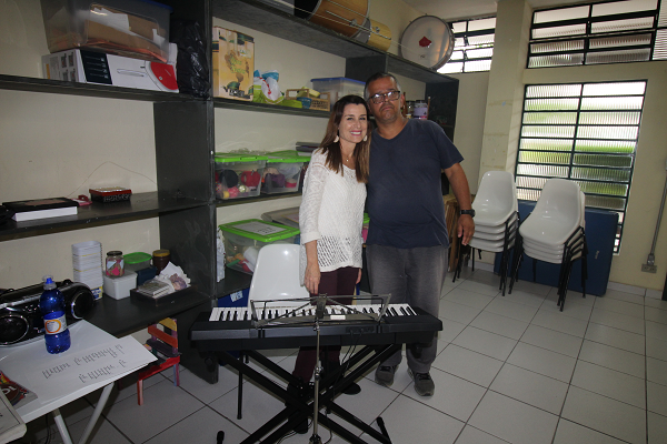 PraCegoVer: A fonoaudióloga e musicista, Cecília Crepaldi abraça o paciente Roberto Silveira. À frente deles está o teclado musical.