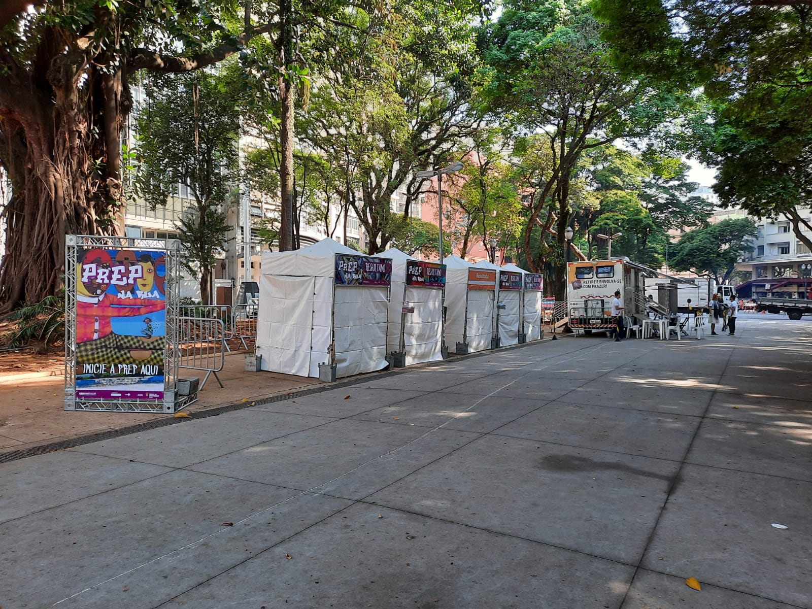 Foto de ambiente. Em um espaço com árvores em volta estão montadas cinco tendas brancas uma ao lado da outra. À esquerda da imagem, está um cartaz colorido com a frase "PrEP na Rua".