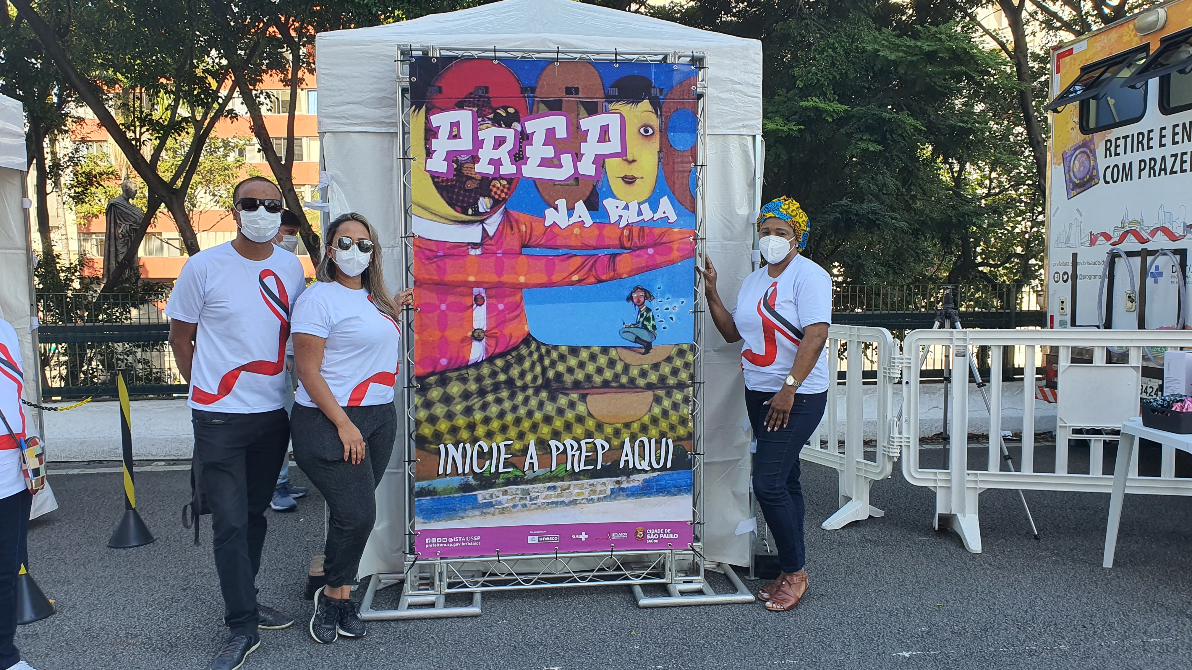Duas mulheres e um homem vestindo camisas brancas com estampa de um laço vermelho. Eles estão ao lado de um cartaz colorido com a frase 