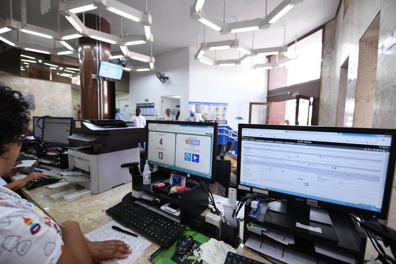 #PraCegoVer: a foto registra a mesa da recepção da UBS República, onde é possível visualizar duas telas de computadores. Em uma das telas há o registro da fila de espera e em outra, o formulário do prontuário eletrônico em exibição.