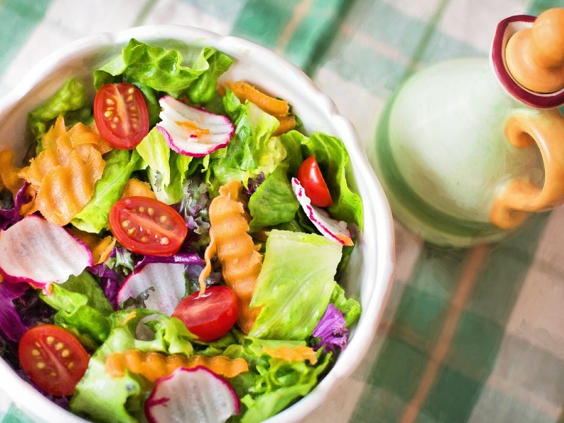Salada colorida posta numa tigela branca, com folhas verdes, tomate, cenoura, rabanete e repolho roxo. Ao lado, um galheteiro