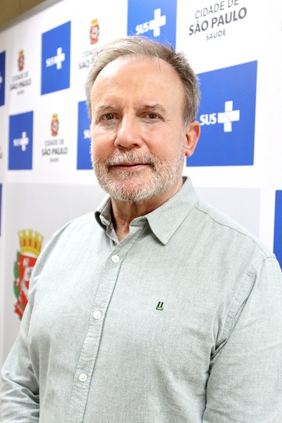 Doutor Zamarco tem cabelo e barba grisalha e está em pé, usando uma camisa clara. Na parede ao fundo os logos dos SUS e da Secretaria Municipal da Saúde