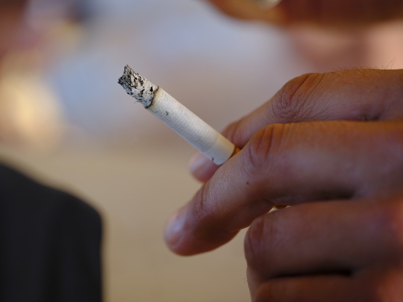 Na foto aparece a mão de uma pessoa segurando um cigarro aceso entre os dedos. O fundo da imagem está desfocado