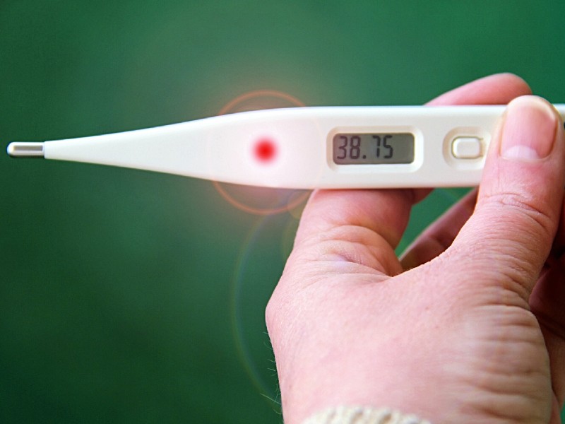 Foto de um termômetro digital sobre um fundo verde. A foto foca na mão de uma pessoa segurando um termômetro branco com a ponta metálica. O visor marca a temperatura de 38,75 graus