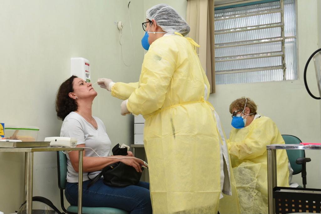 Uma profissional de saúde vestindo jaleco amarelo, touca branca e máscara de proteção azul, de óculos, está coletando o teste de Covid-19 das narinas de uma mulher, que está sentada em uma cadeira de encosto verde. Ela veste camisa branca e calça jeans azul. Atrás delas, está outra profissional de saúde sentada.