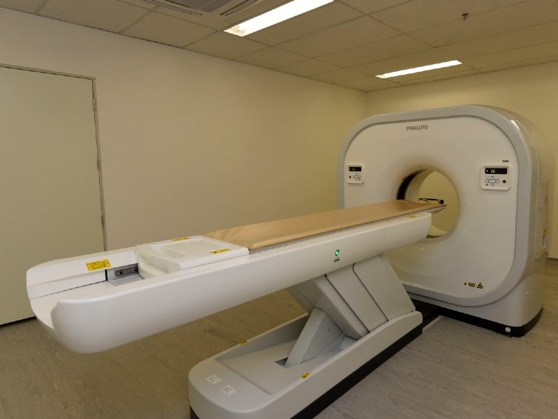 Foto do tomógrafo do Hospital Lydia Storópoli. O equipamento é composto por uma plataforma onde o paciente fica deitado e o aparelho que realiza o exame, em formato quadrado com uma abertura no meio