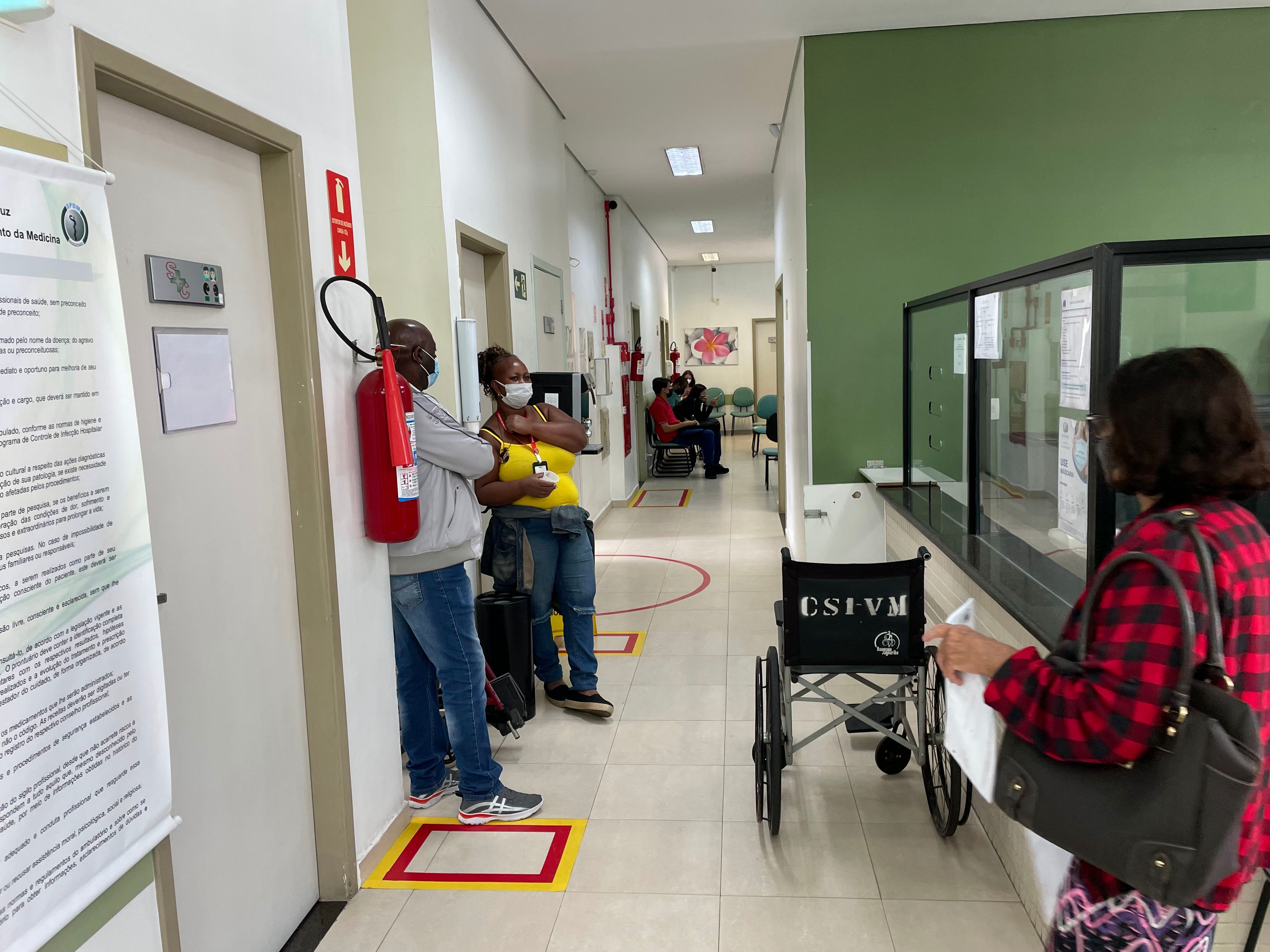 Foto de corredor. No corredor da UBS, estão duas pessoas encostadas em uma das paredes. Em frente à essas pessoas, está uma cadeira de rodas vazia. Ao lado deles, tem uma porta de consultório fechada. No canto direito da imagem, uma mulher está parada de costas.