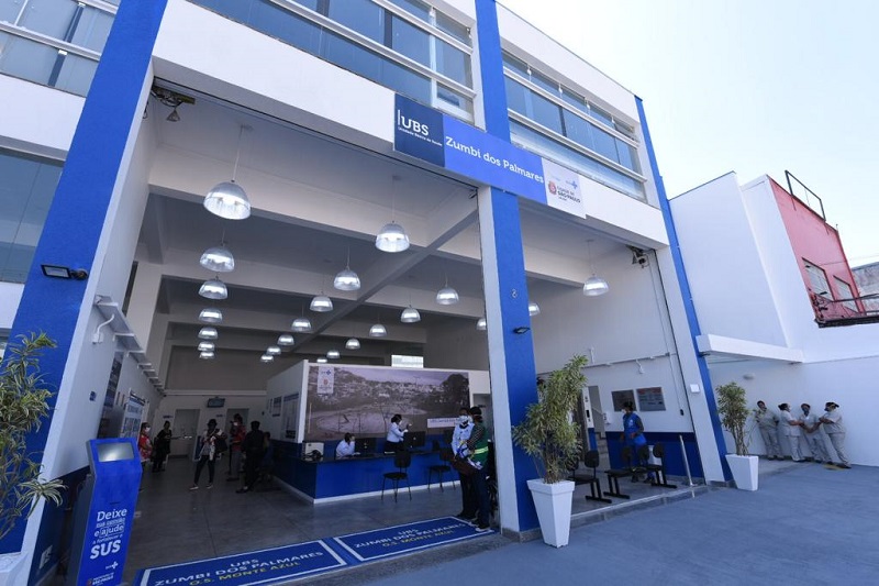 Foto da fachada da UBS Zumbi dos Palmares. A fachada é azul e branca e tem várias luminárias.É possível observar alguns profissionais de saúde e munícipes.