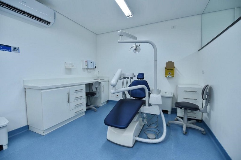 Foto do consultório de odontologia. No centro há uma cadeira odontológica e à esquerda há um móvel branco com gavetas.