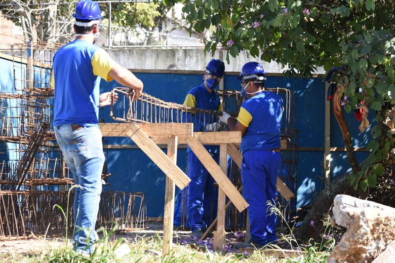 #PraCegoVer: três profissionais da construção civil, com uniformes azuis e equipamentos de proteção individual, trabalham na obra da UPA Maria Antonieta.