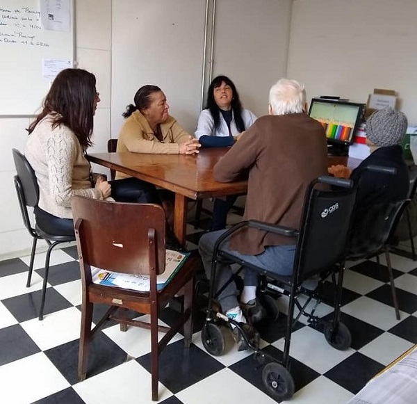 #PraCegoVer: na imagem há quatro idosos, sendo três mulheres e um homem, e uma profissional de saúde. Eles estão sentados em volta de uma mesa de madeira.