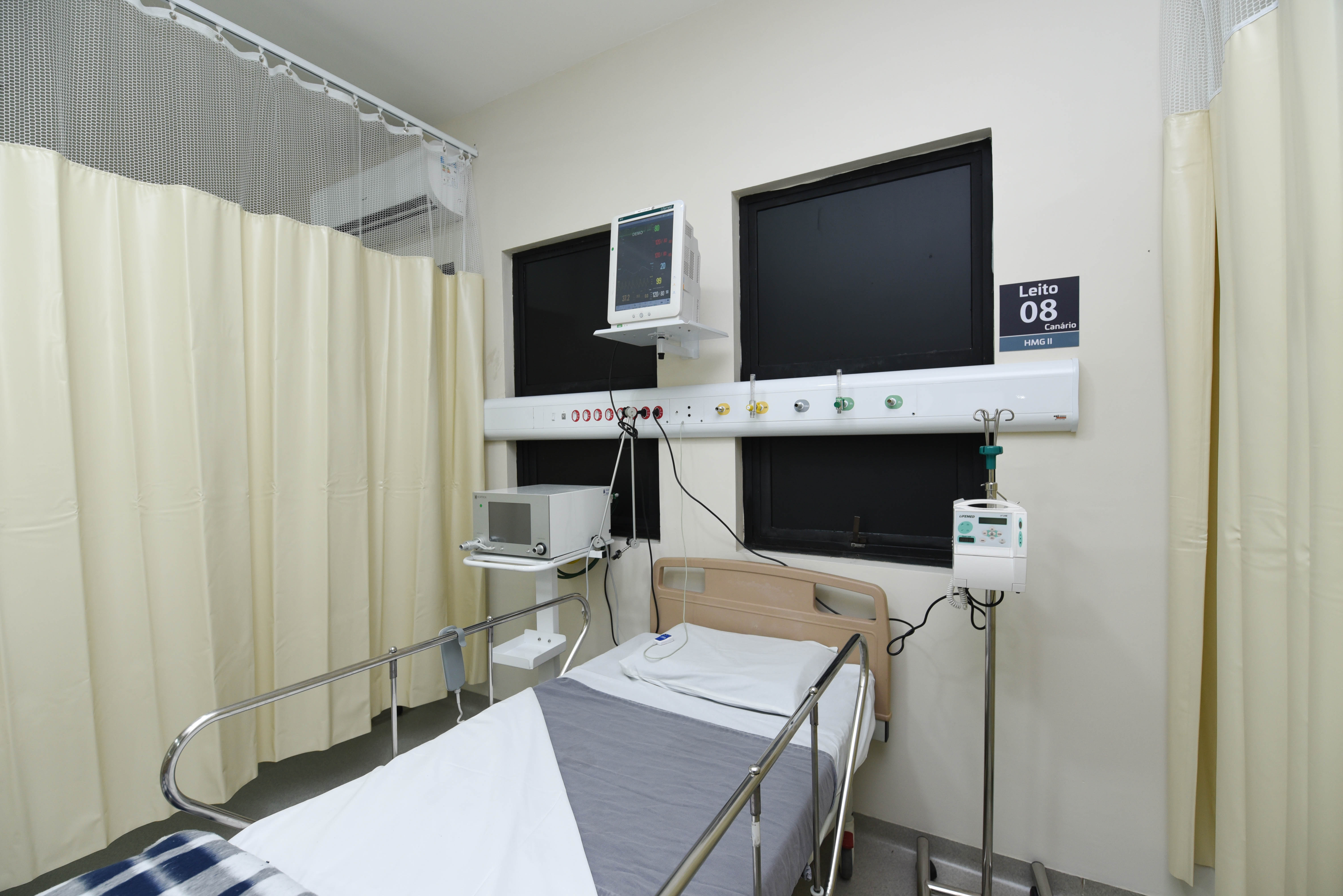 Foto de UTI. Uma cama de hospital está no centro do cômodo. Na parede em que ela está encostada estão duas janelas e uma plaquinha quadrada com 